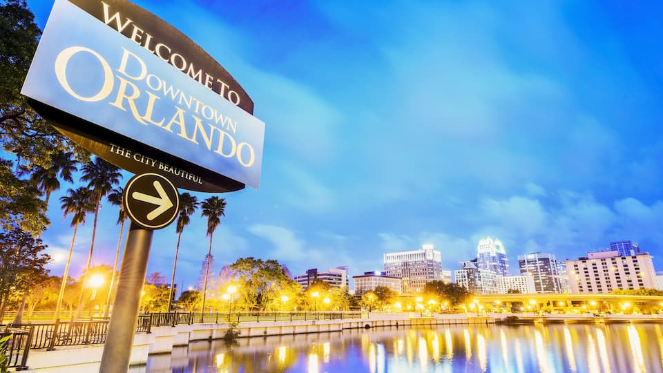 A sign reading "Welcome to Downtown Orlando" with a reflection in the water and city skyline in the background.