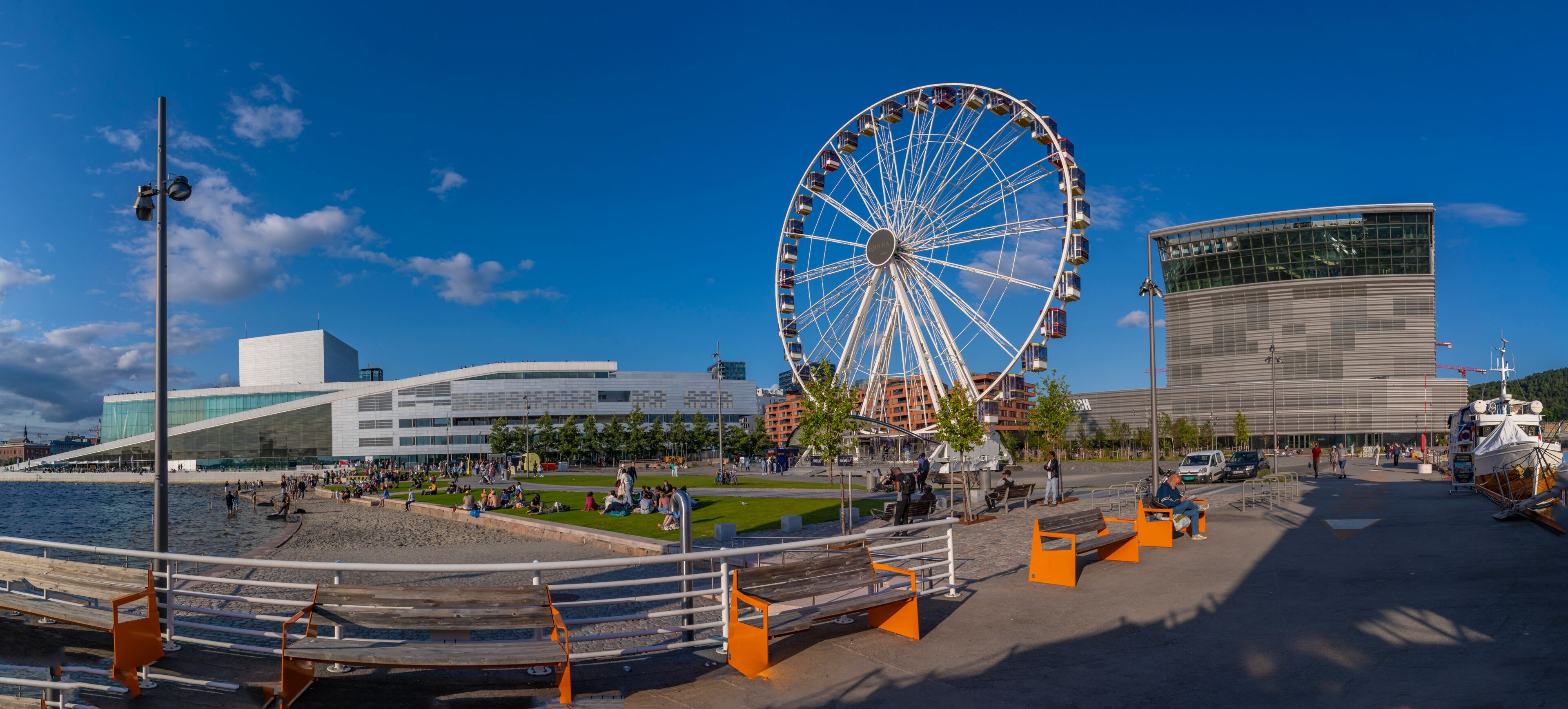 A waterfront promenade with a large Ferris wheel, modern buildings, and a clear blue sky.