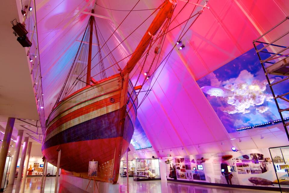 A large ship suspended in a modern building with colorful lighting and a cloud mural on the wall.