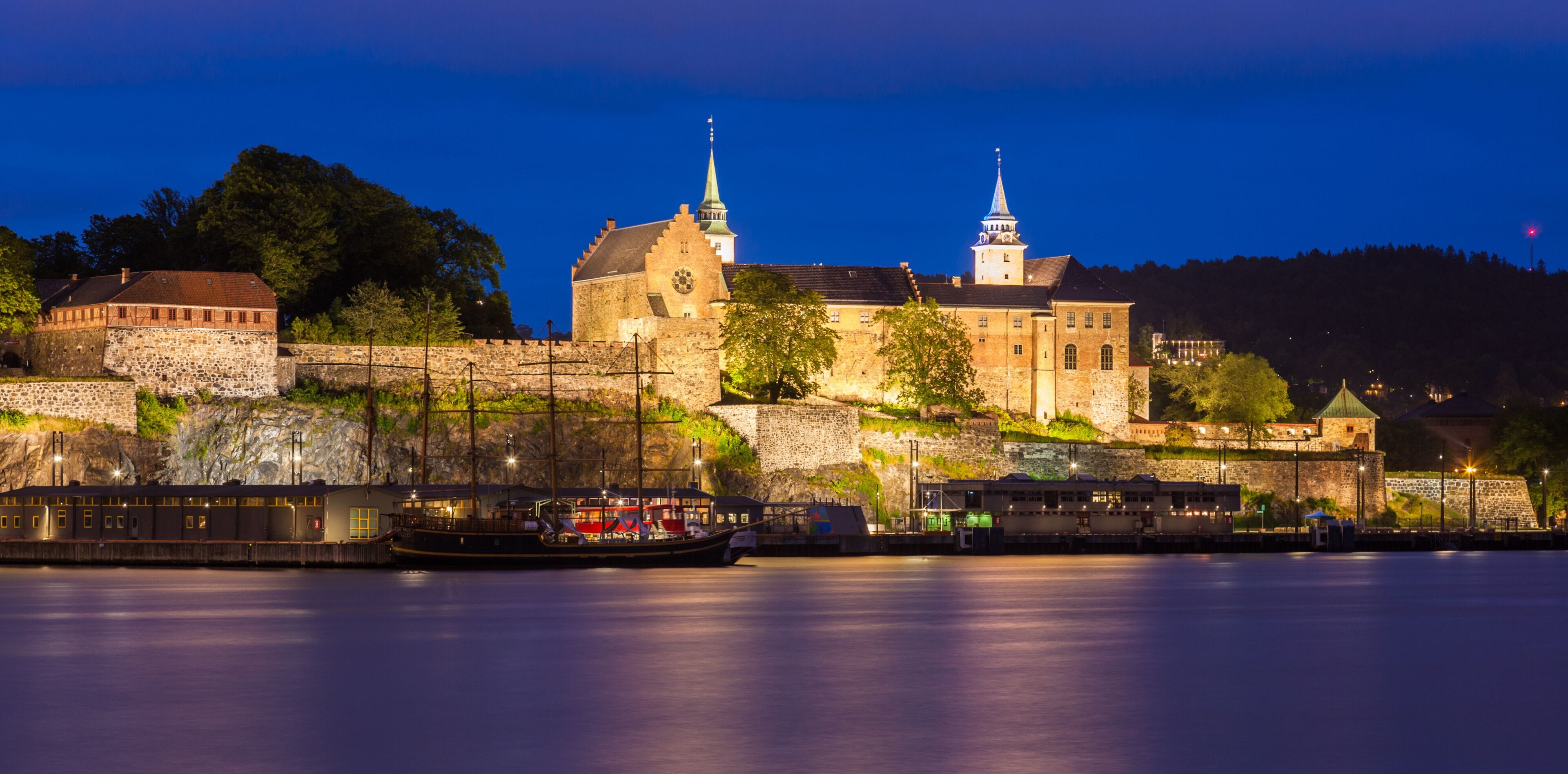 A historic castle with two spires, a river, and a boat docked at the pier.