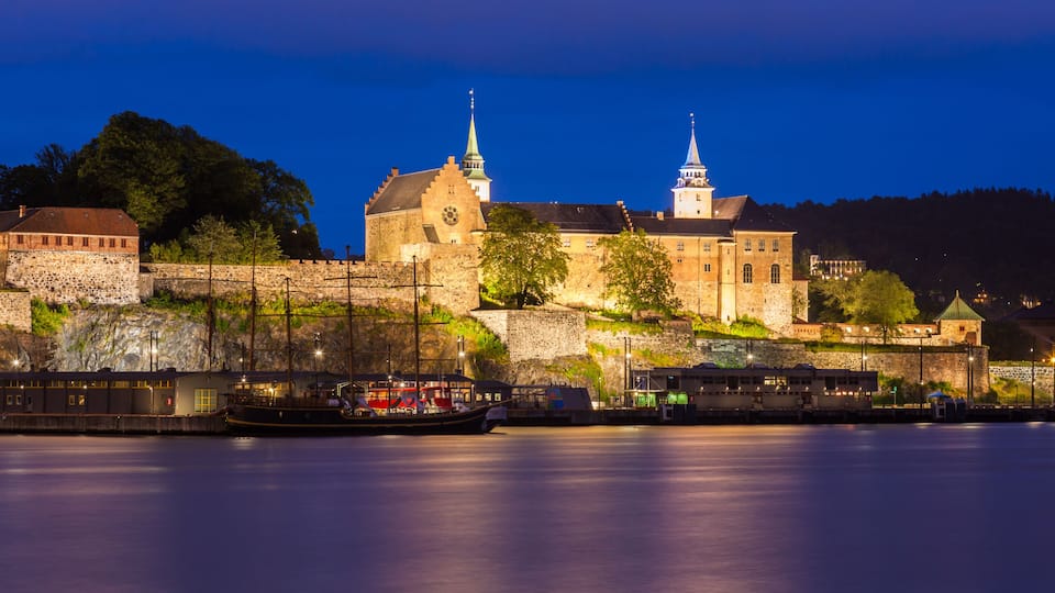 A historic castle with two spires, a river, and a boat docked at the pier.