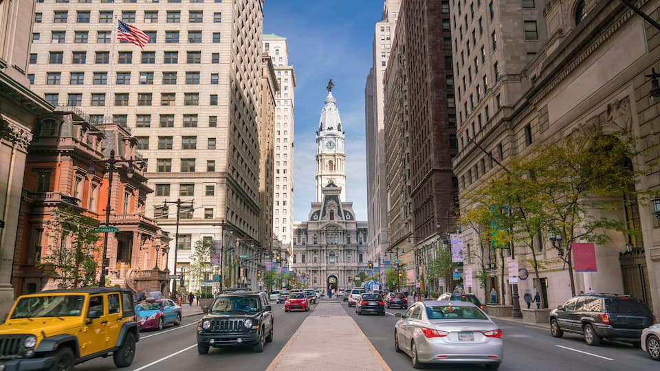 A city street with tall buildings, a prominent clock tower, and cars driving on the road.