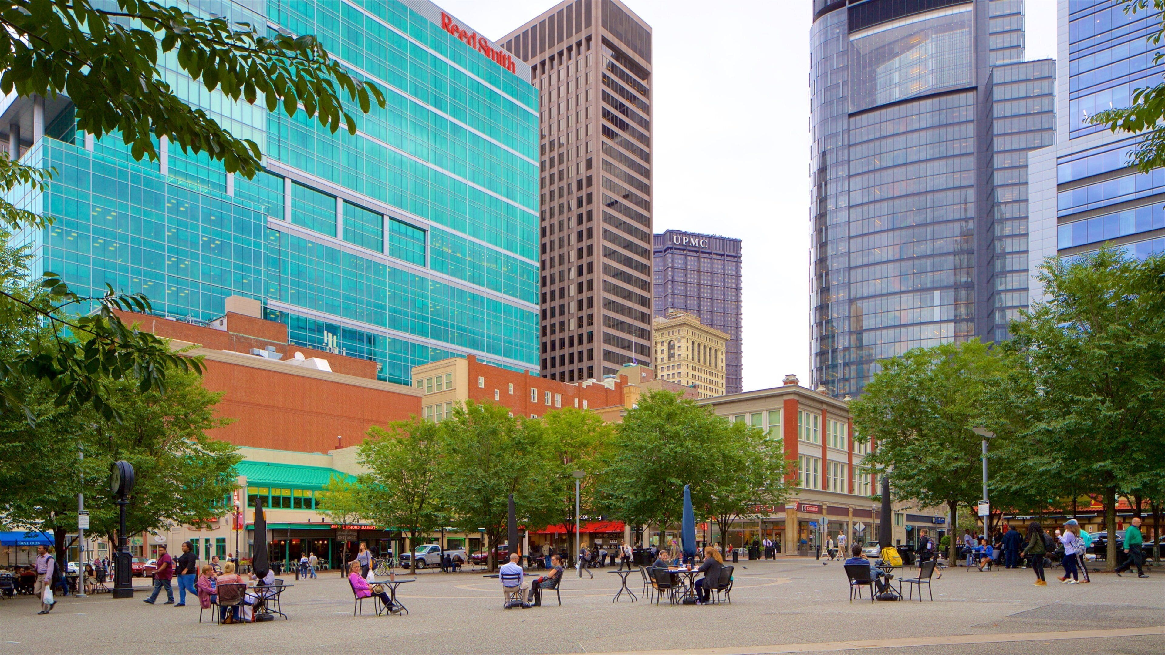 Una plaza urbana con edificios modernos, árboles y gente sentada en mesas al aire libre.