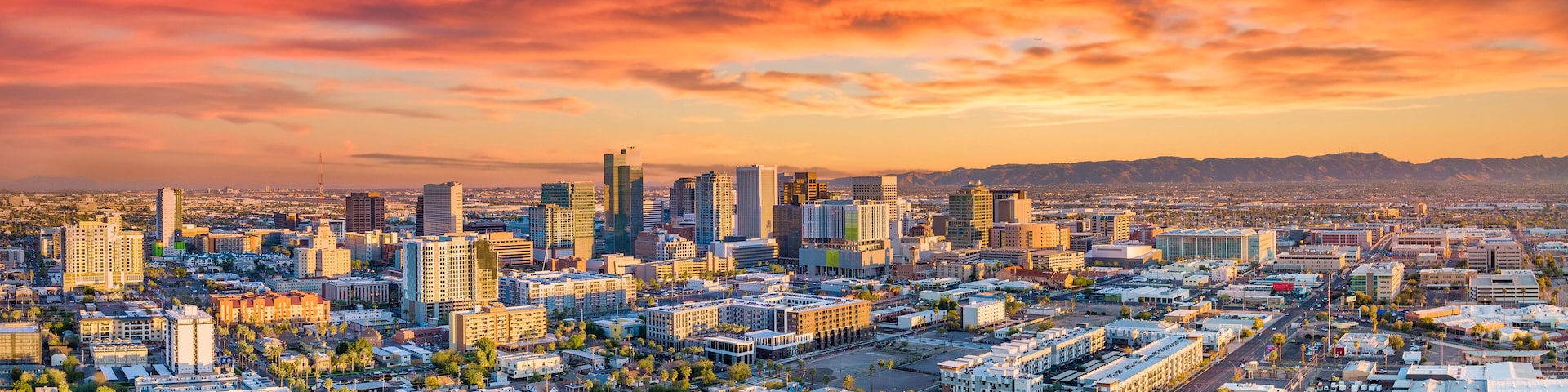Paisagem urbana com céu ao pôr do sol, edifícios e uma cadeia de montanhas ao longe.