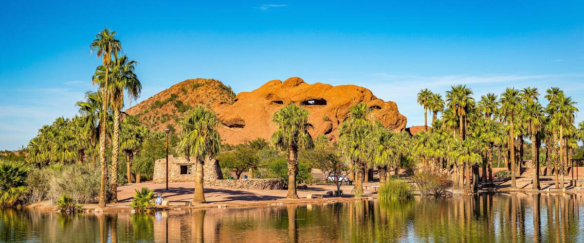 A desert landscape with a large rock formation, palm trees, and a body of water reflecting the scenery.