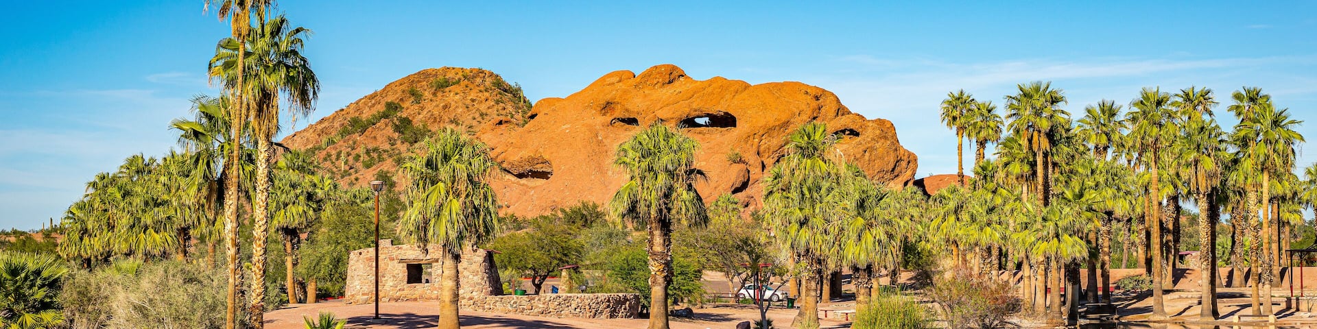 A desert landscape with a large rock formation, palm trees, and a body of water reflecting the scenery.