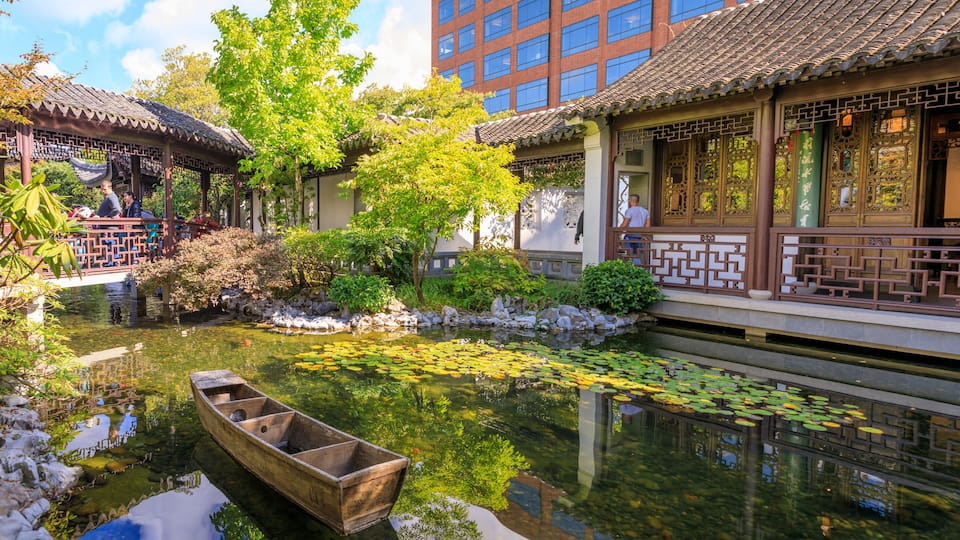 A traditional Chinese garden with a wooden boat, a pond with lily pads, and ornate buildings.