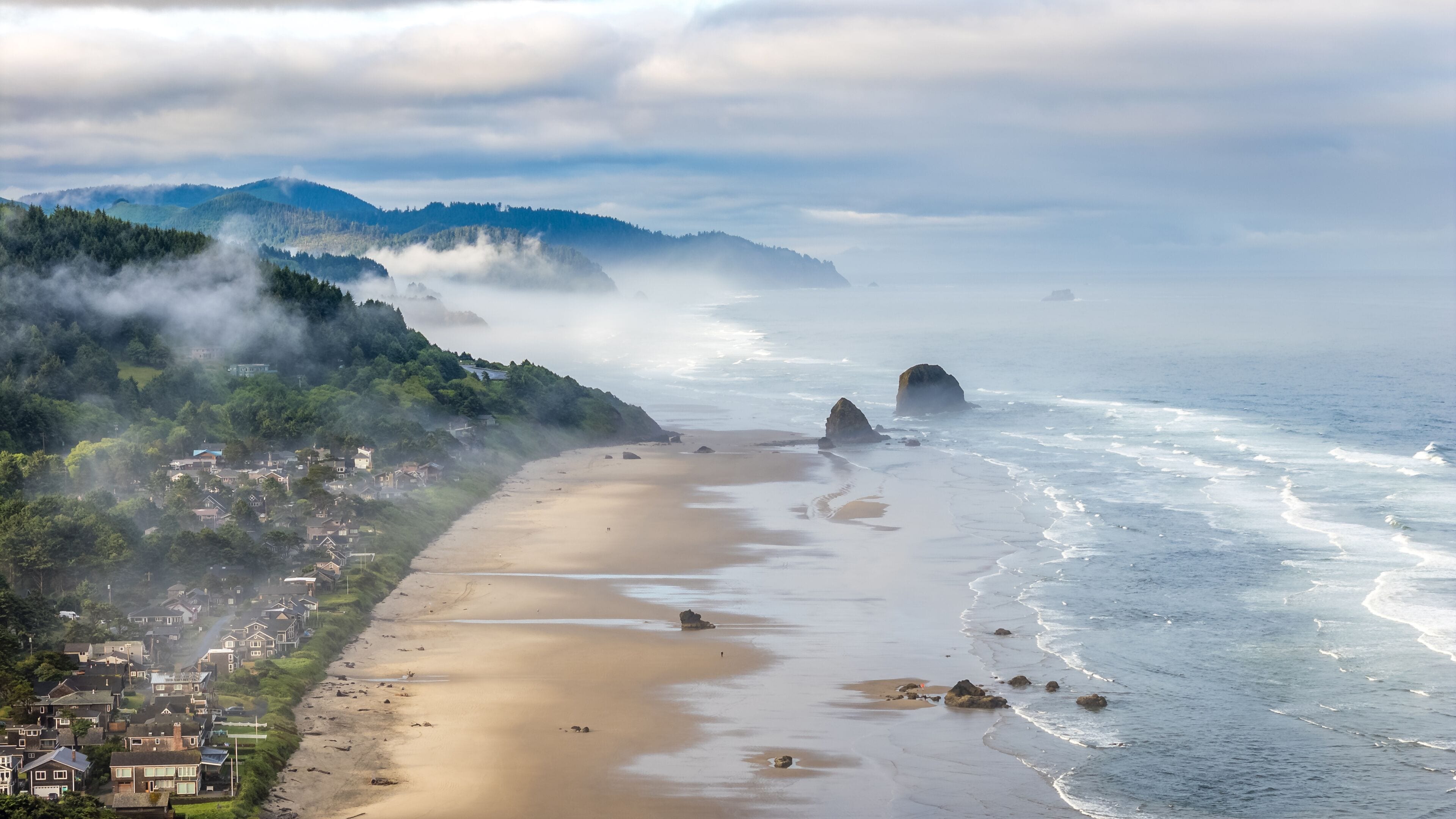 A coastal town with a sandy beach, rocky outcrops, and forested hills.