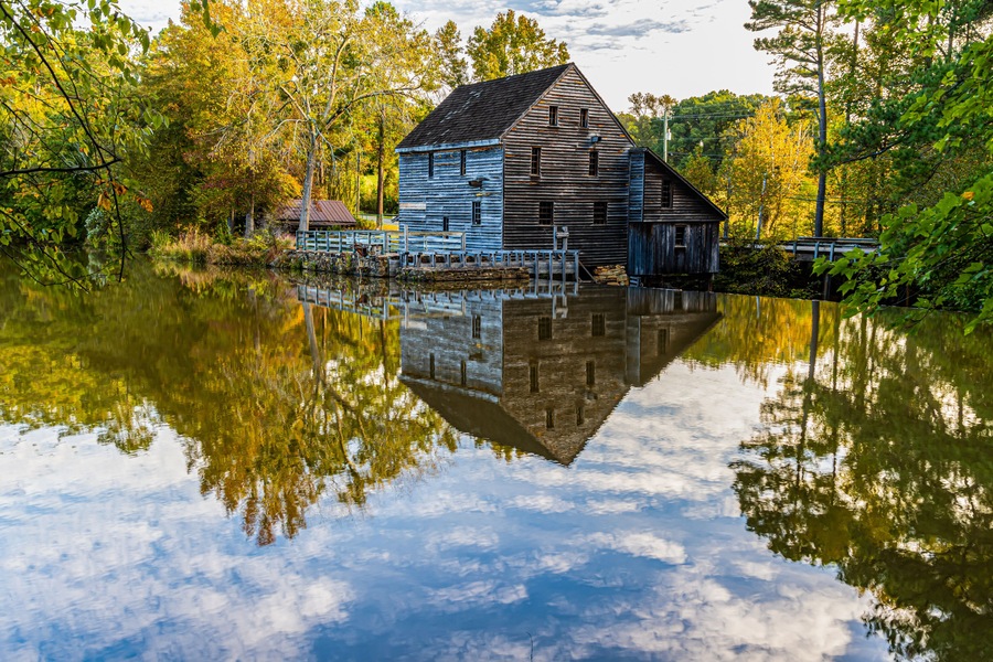 Una casa de madera junto a un lago tranquilo, con reflejos de árboles y cielo.