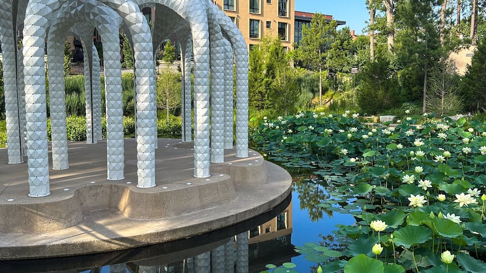 A modern sculpture with geometric shapes, a pond with lily pads, and a building in the background.