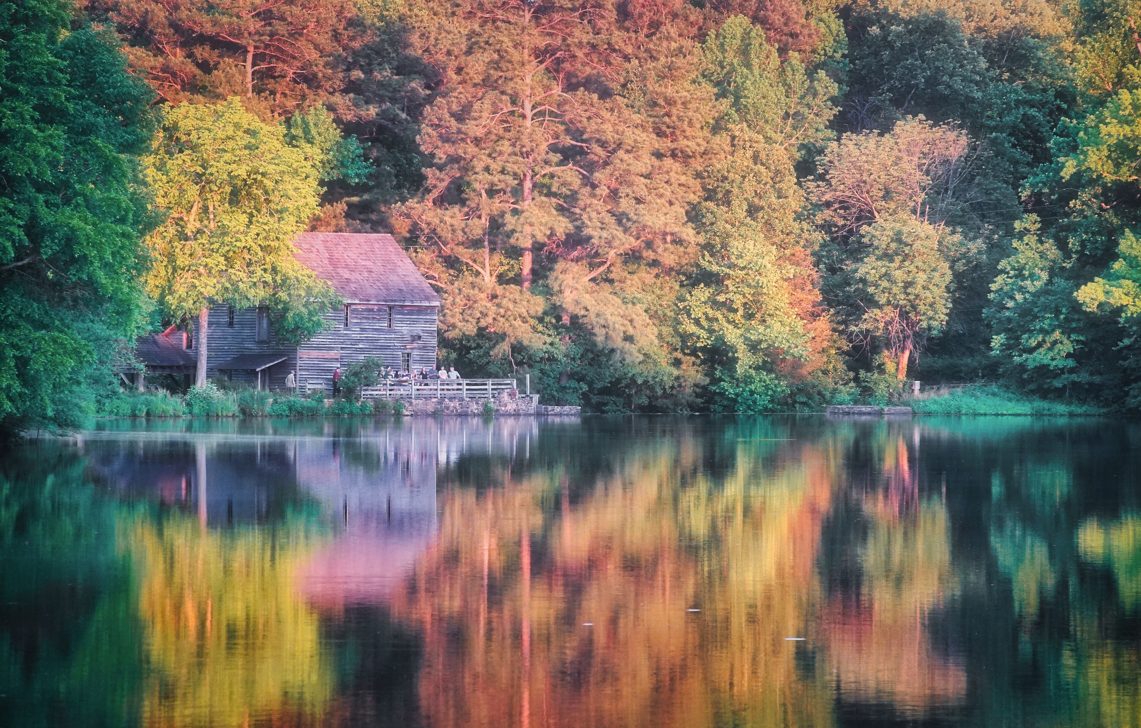 A wooden cabin by a calm lake surrounded by autumn-colored trees.