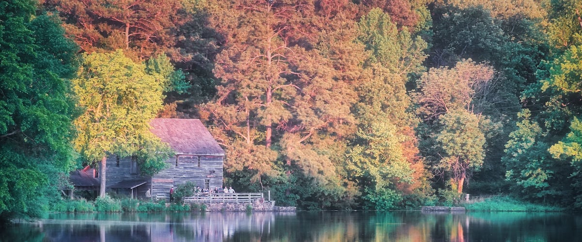 A wooden cabin by a calm lake surrounded by autumn-colored trees.