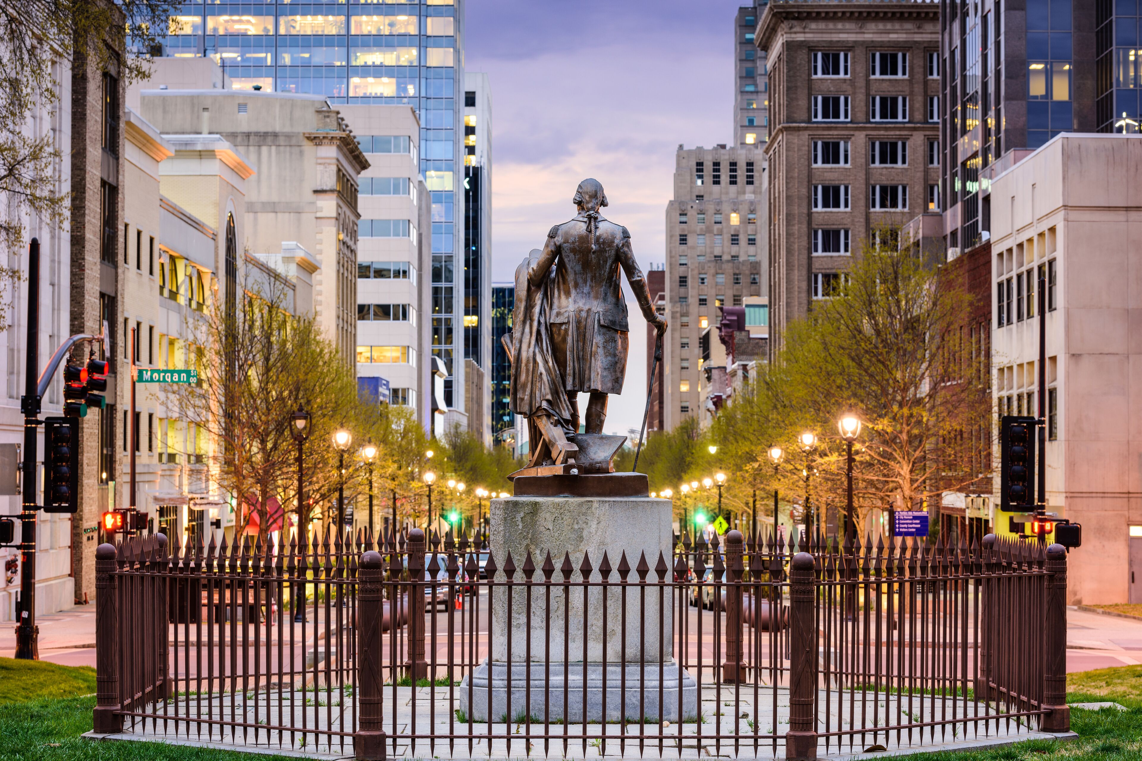 A statue in the center of a city square surrounded by a fence, with buildings and trees in the background.
