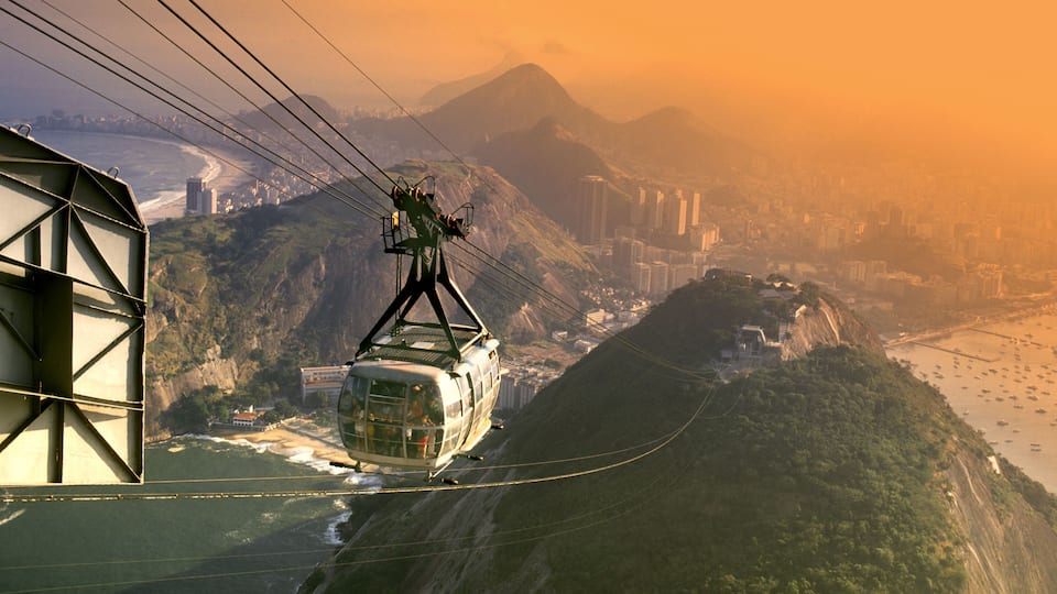 A cable car moving along a cable over a mountainous landscape with a city in the background.