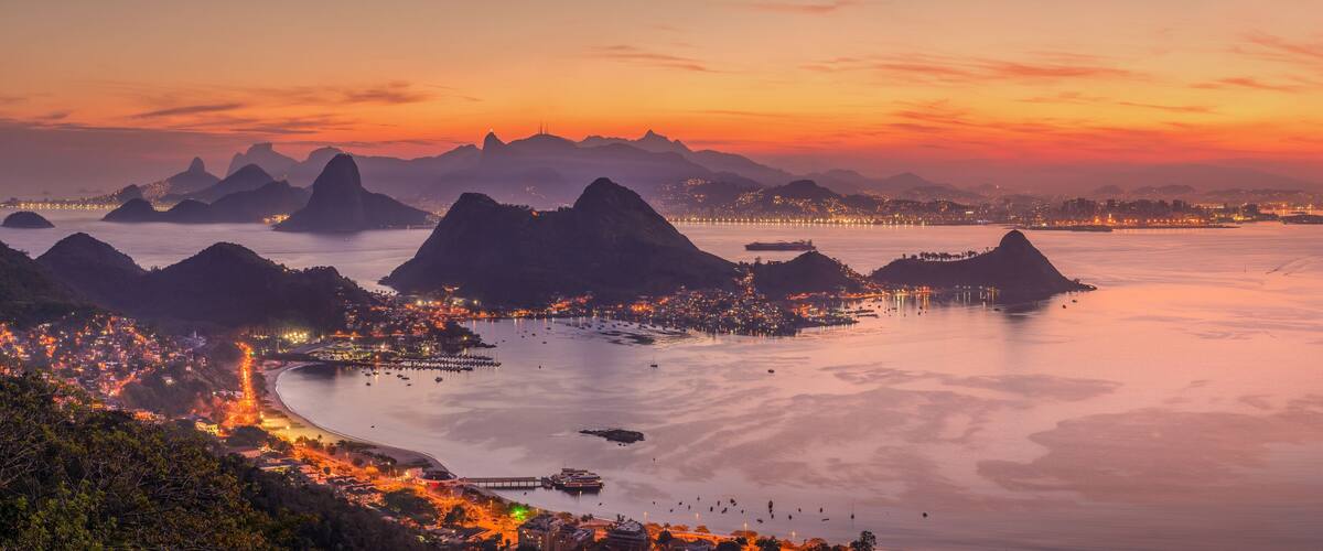A coastal cityscape at sunset with illuminated buildings, a calm bay, and prominent mountain ranges.
