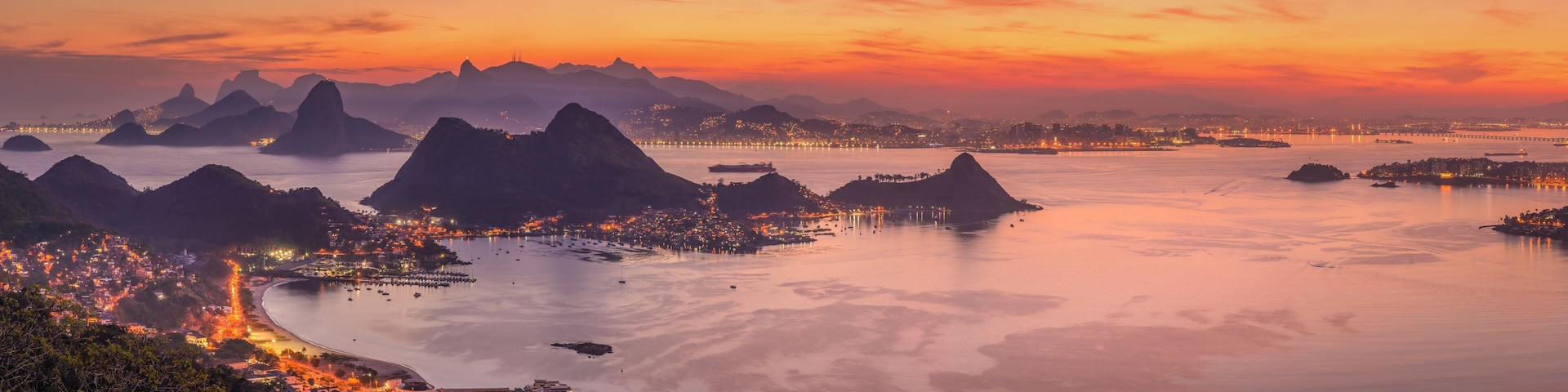 A coastal cityscape at sunset with illuminated buildings, a calm bay, and prominent mountain ranges.