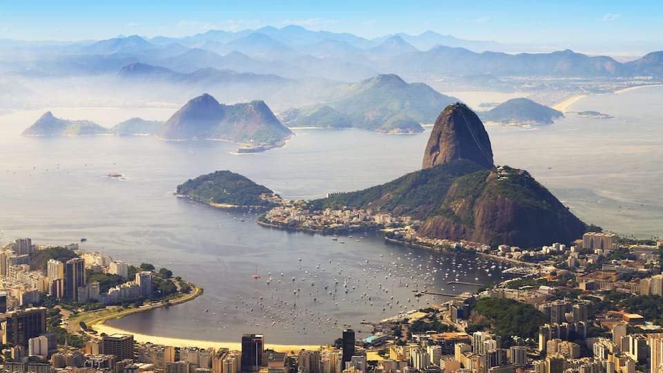 A coastal cityscape with prominent mountains, a bay filled with boats, and dense urban buildings.
