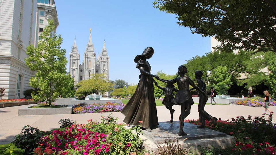 A statue of a woman and child in a park with flowers and a cathedral in the background.