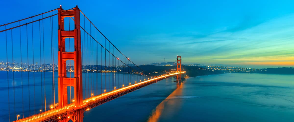 A suspension bridge with illuminated cables, a calm body of water, and a distant city skyline at dusk.