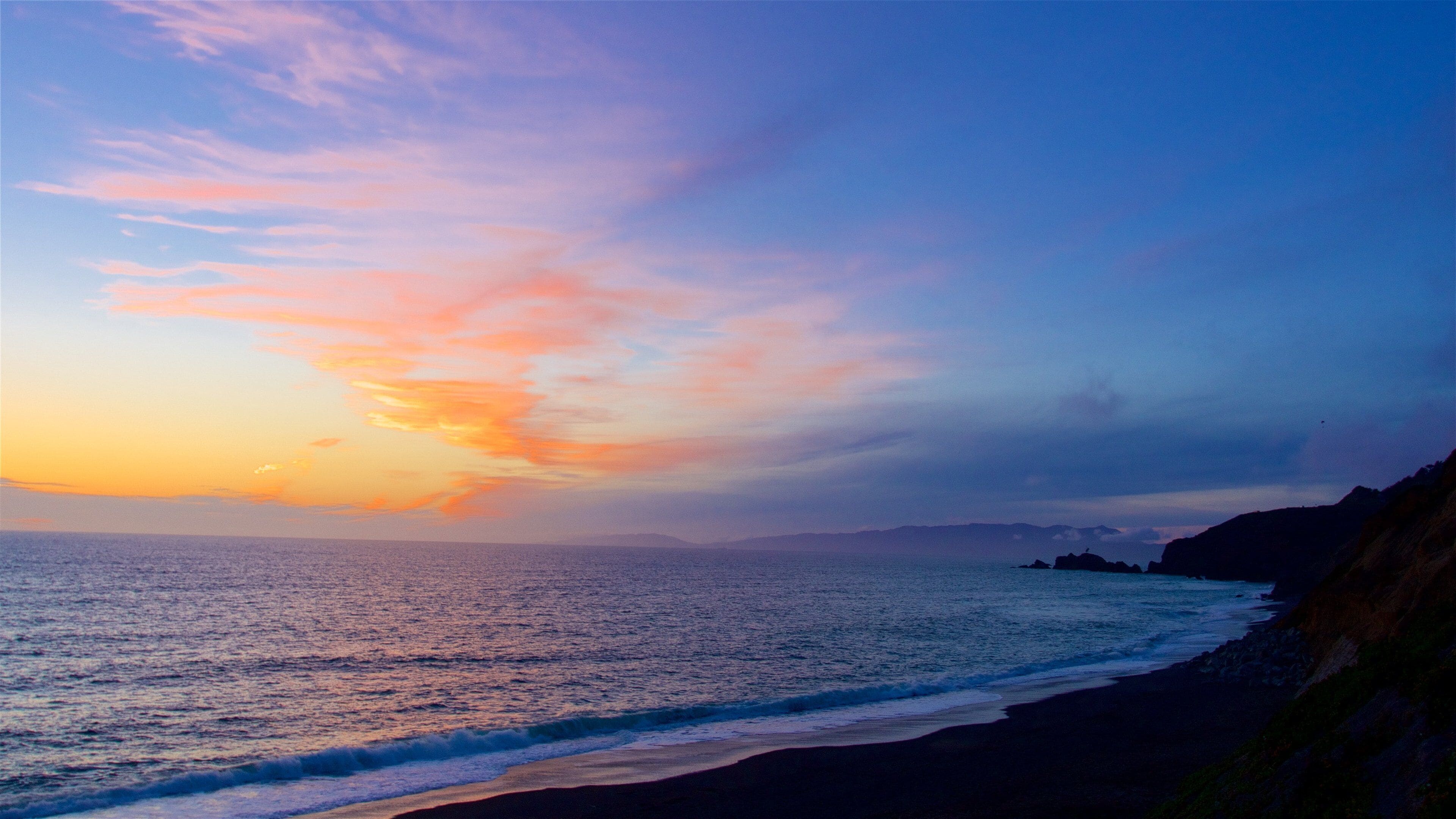 A sunset over a coastal landscape with a beach and rocky cliffs.