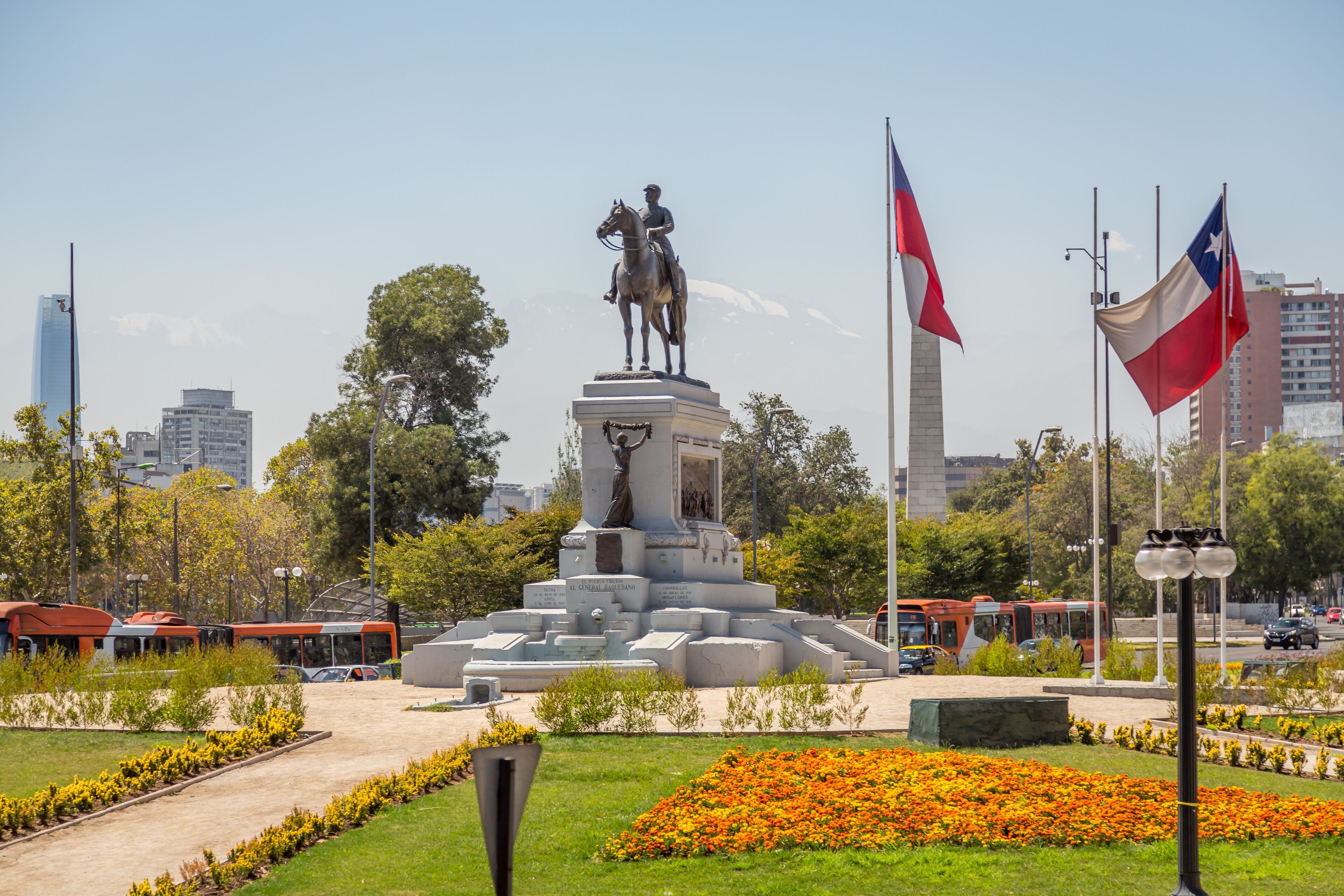 A statue of a man on horseback, surrounded by a landscaped garden with orange flowers, set against a cityscape with tall buildings and flags.