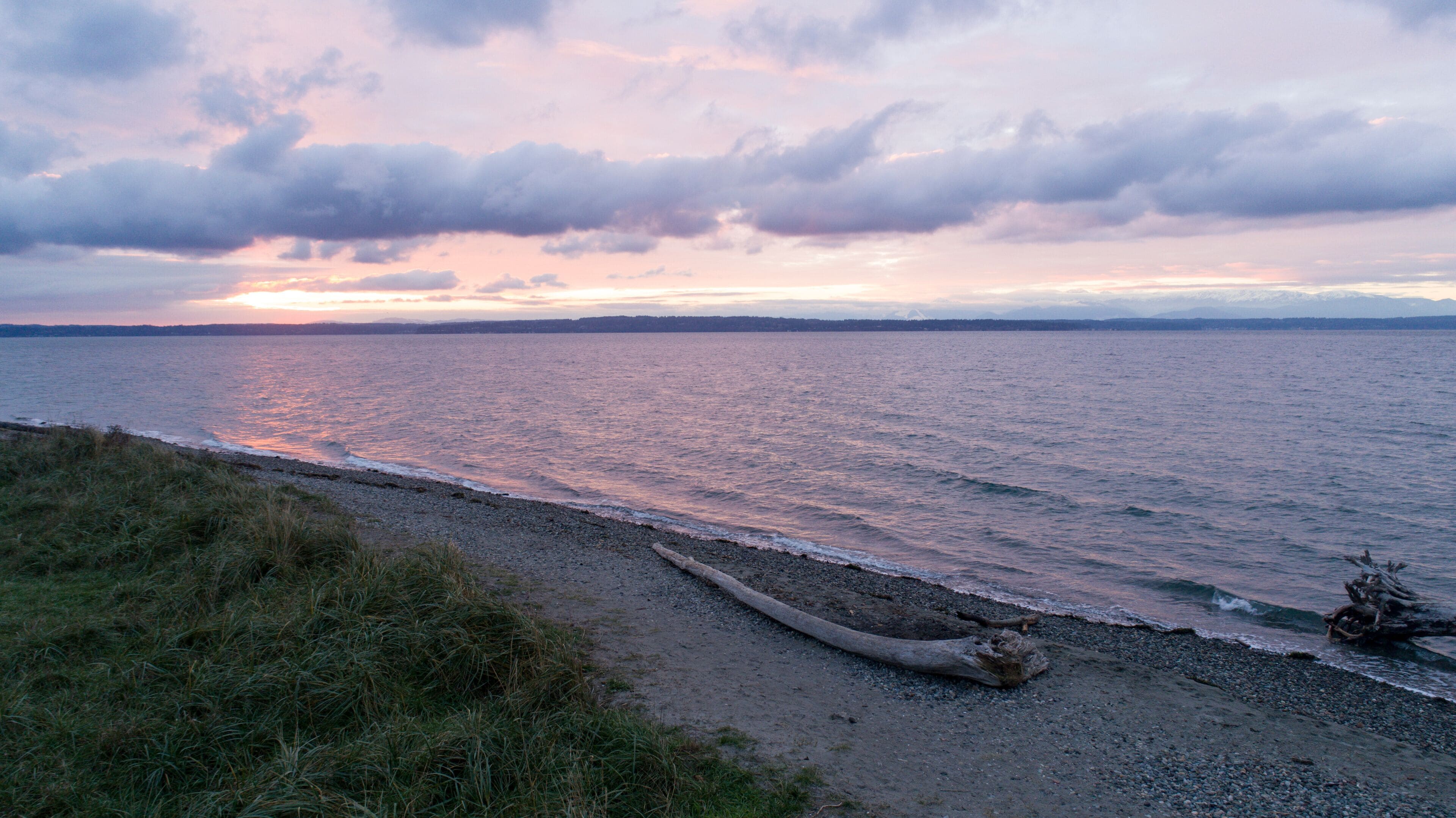 A coastal landscape with a pebbly beach, a fallen tree, and a sunset over the water.