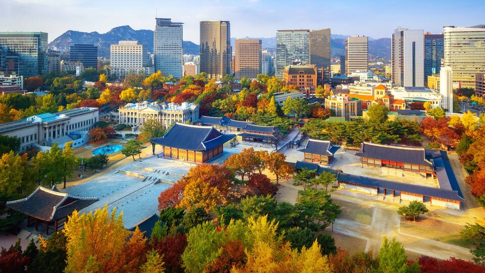 A cityscape with traditional architecture amidst modern skyscrapers, autumn foliage, and a mountainous backdrop.