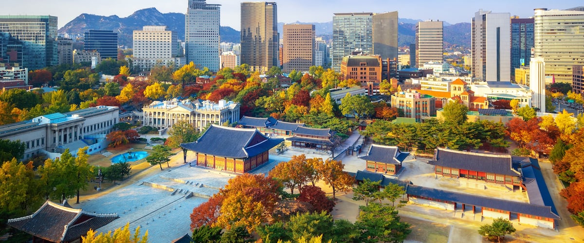 A cityscape with traditional architecture amidst modern skyscrapers, autumn foliage, and a mountainous backdrop.