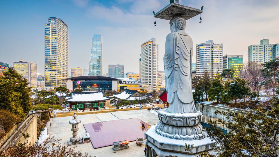 A large statue in a park with traditional architecture and modern skyscrapers in the background.