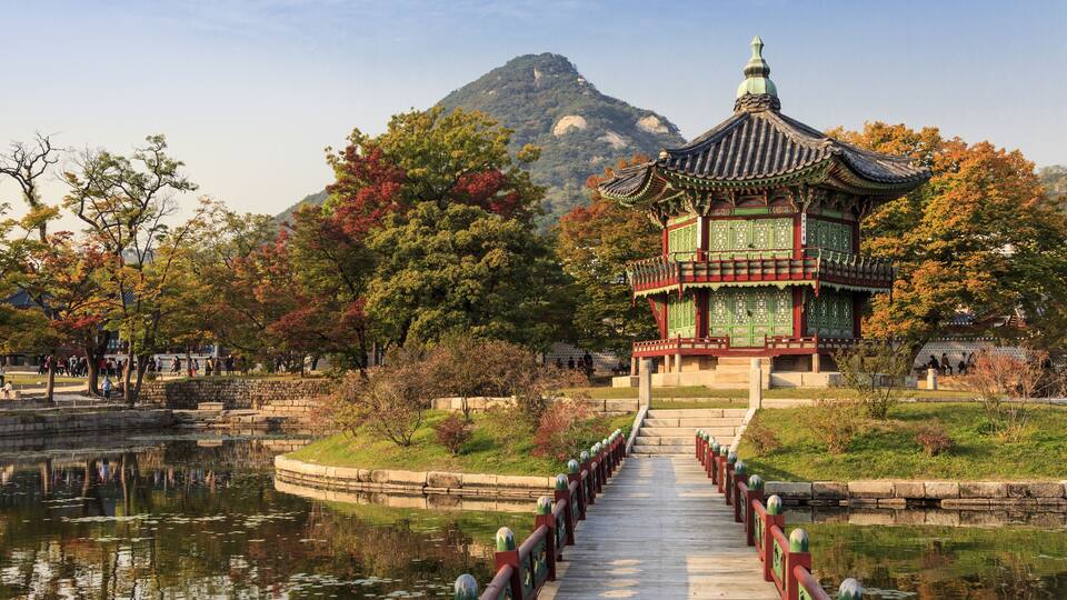 A traditional pavilion with a tiled roof, a wooden walkway leading to it, and a mountain in the background.