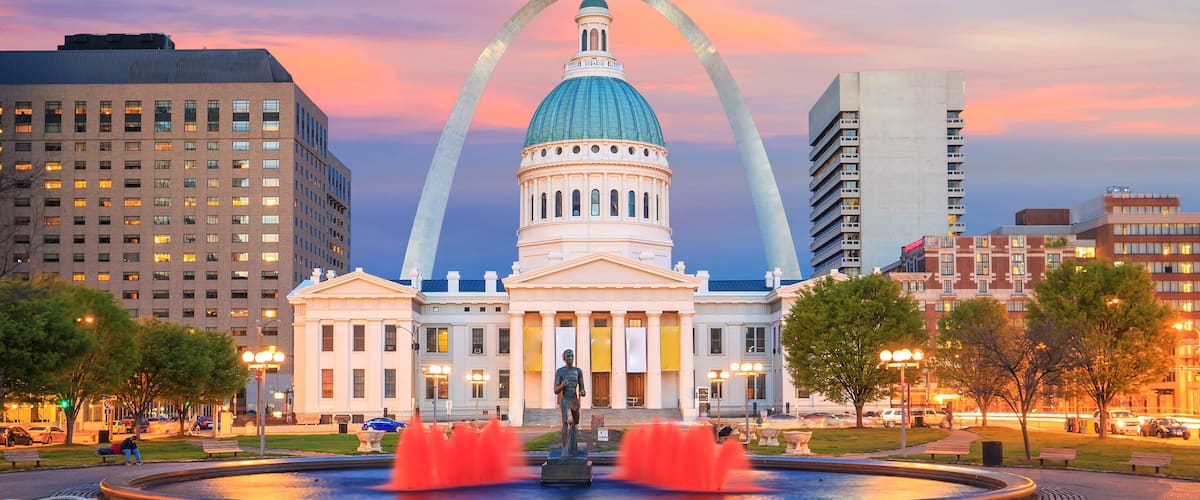 A large white building with a dome and a statue in front, surrounded by fountains and trees.