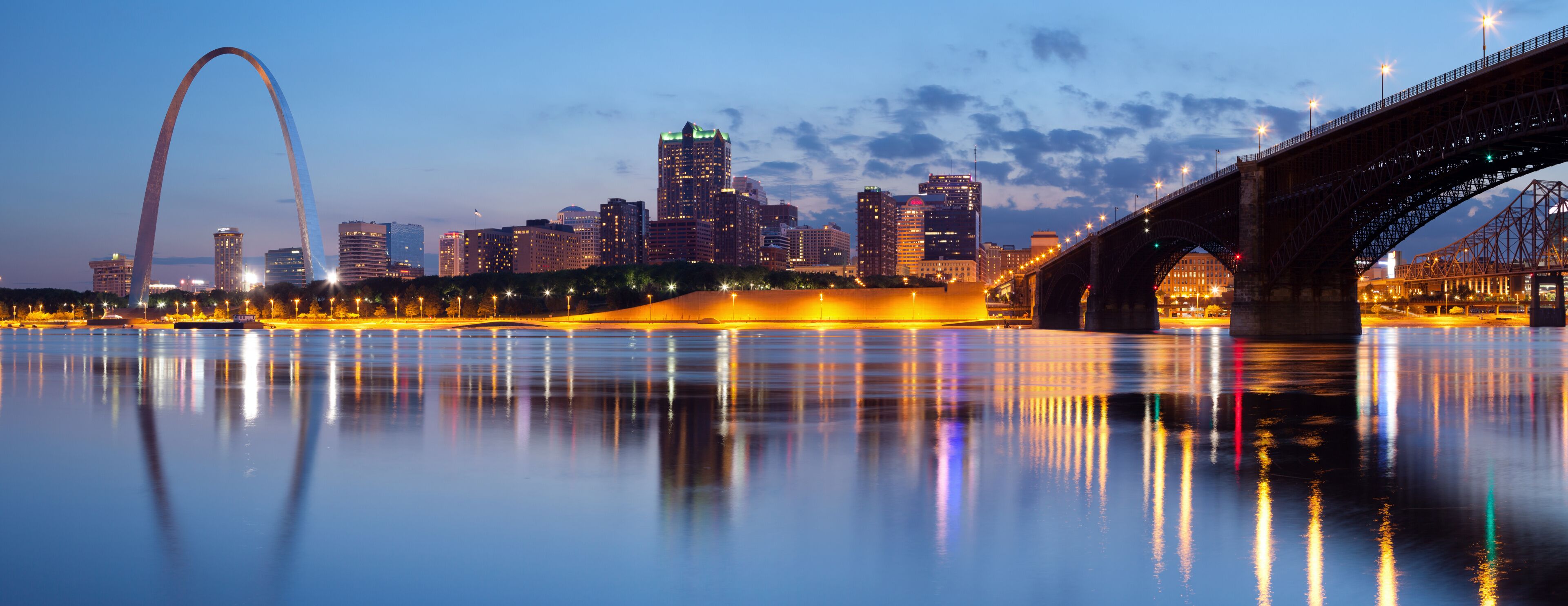 A city skyline with a prominent arch bridge and a lit bridge over a calm river reflecting city lights.