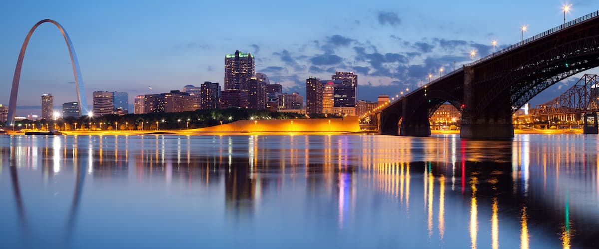 A city skyline with a prominent arch bridge and a lit bridge over a calm river reflecting city lights.