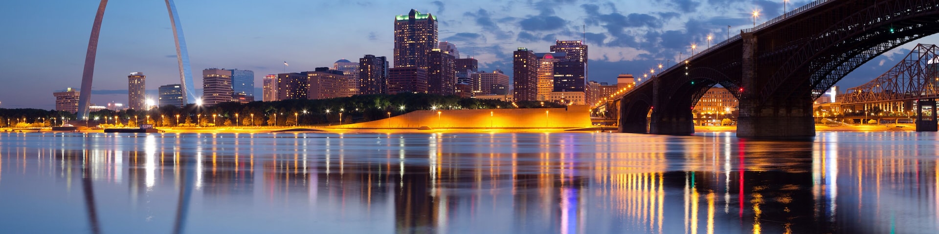 A city skyline with a prominent arch bridge and a lit bridge over a calm river reflecting city lights.