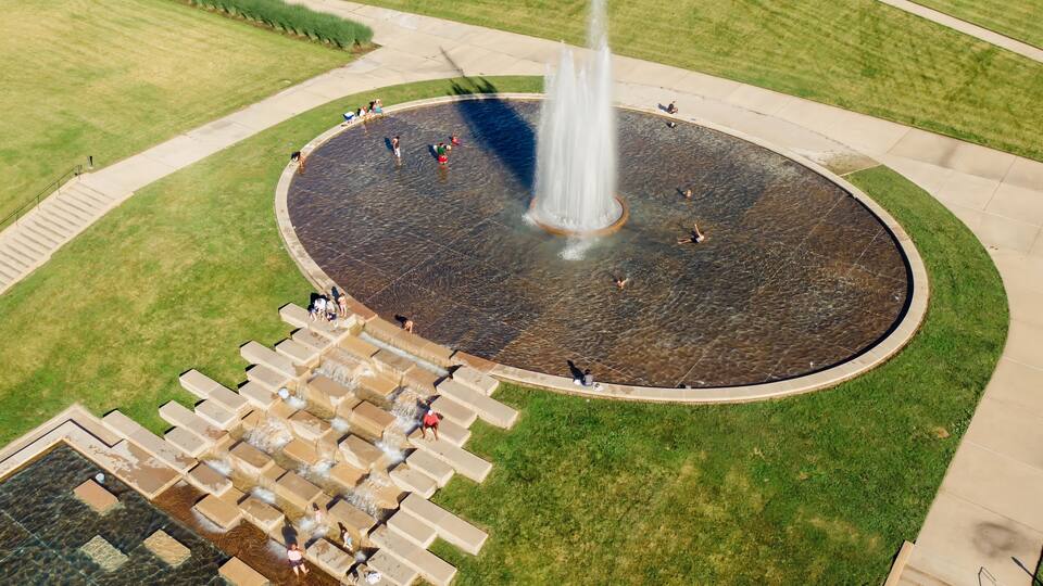 A circular water fountain with a central jet, surrounded by a paved area with stepping stones leading to a pool.
