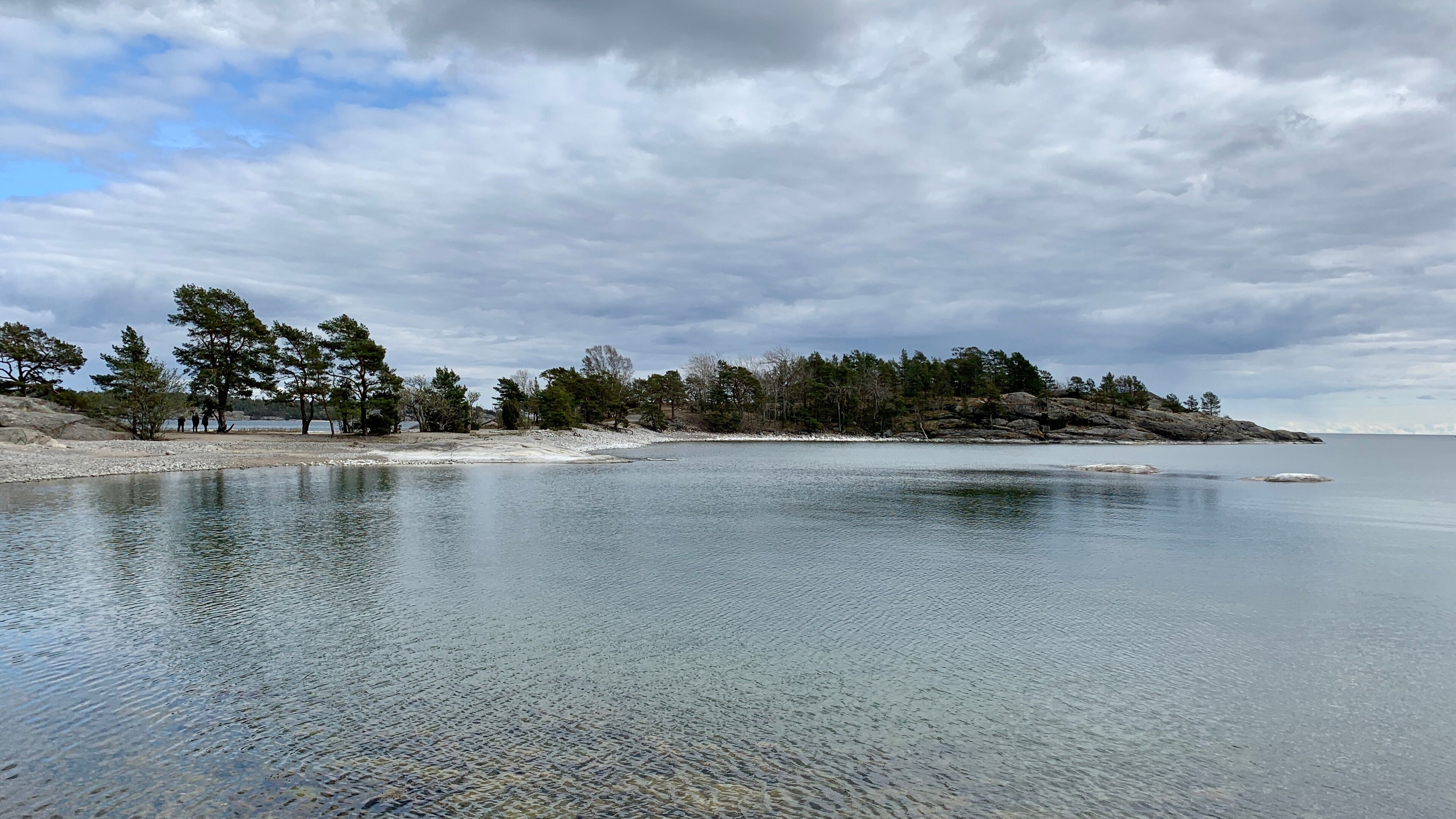 A calm lake with a rocky shoreline, surrounded by trees and a cloudy sky.