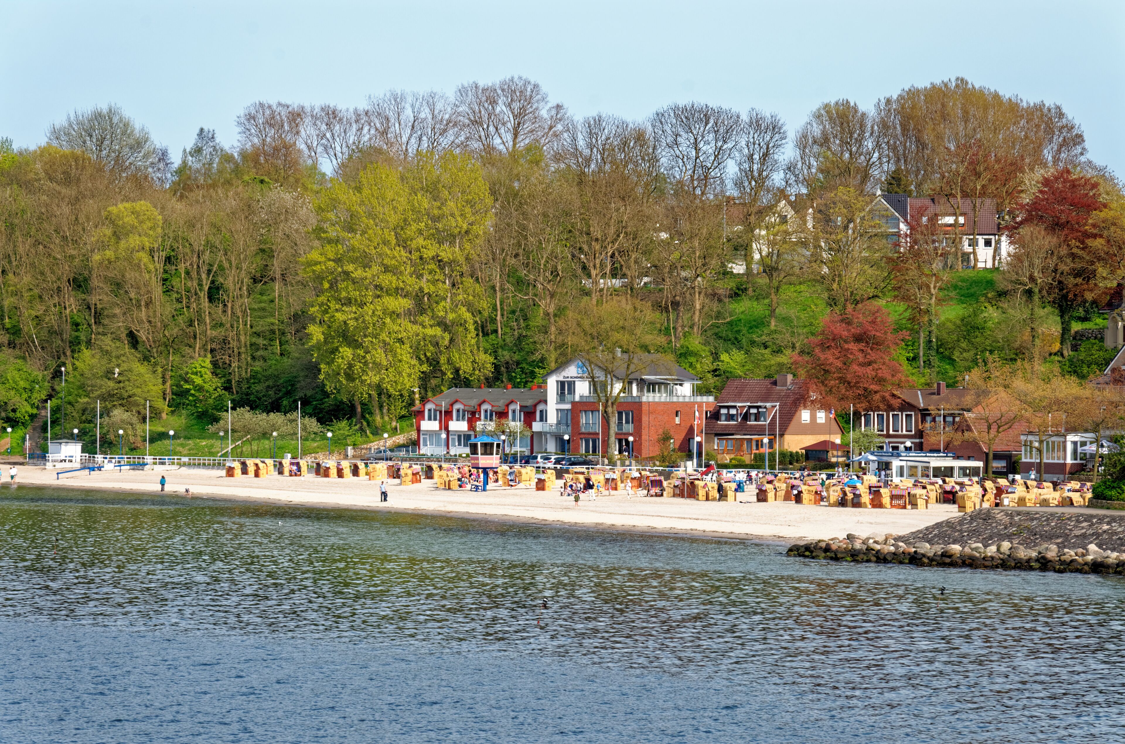 A lakeside beach with wooden beach huts and a row of houses in the background.