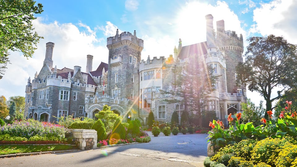 A large, historic stone castle with multiple towers and turrets, surrounded by well-maintained gardens and a paved driveway.
