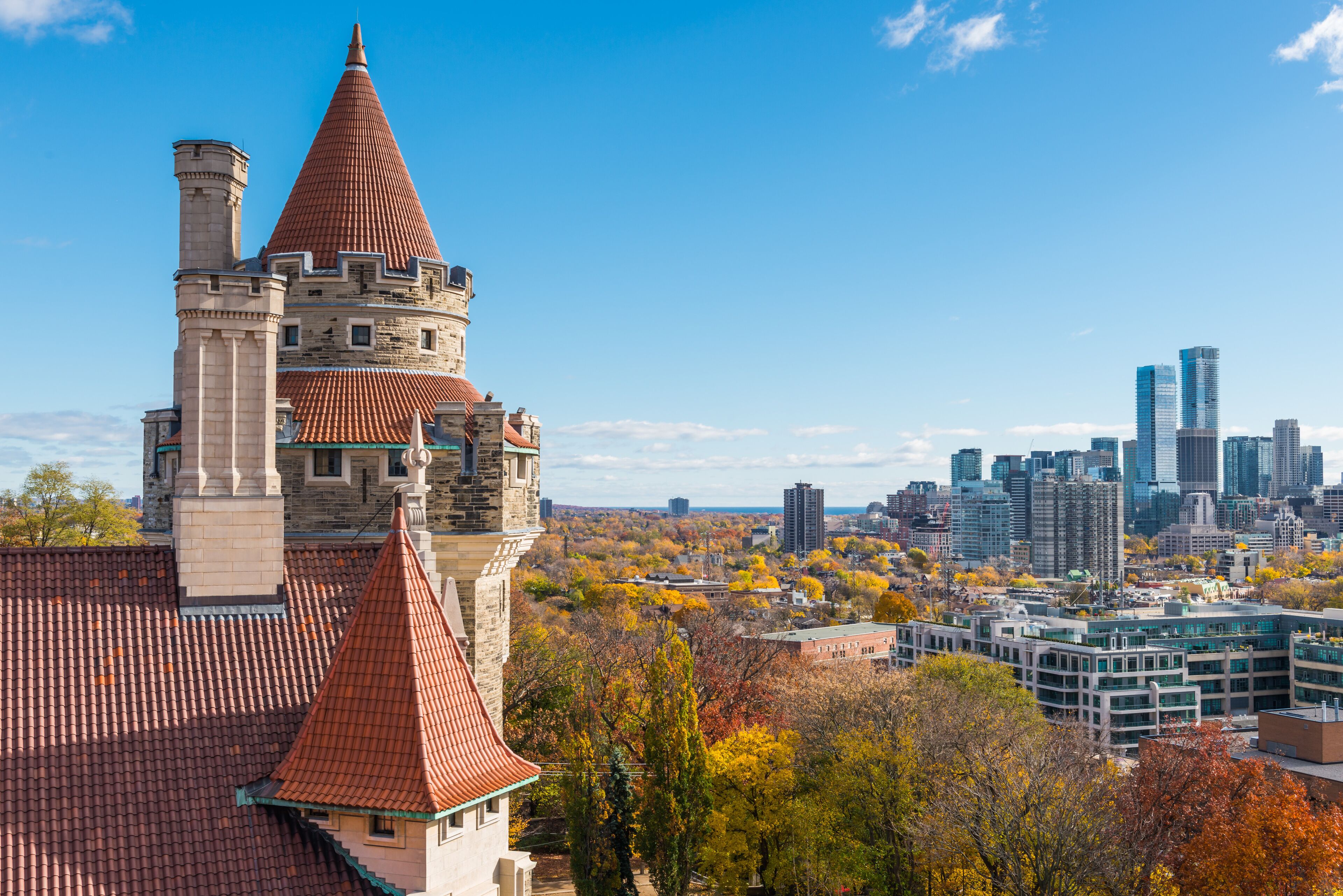 A historic building with a red-tiled roof and a pointed tower, surrounded by autumn foliage and a modern city skyline in the background.