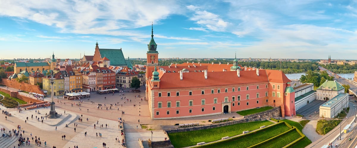 A historic square with a prominent red building, a green-domed tower, and a river in the background.