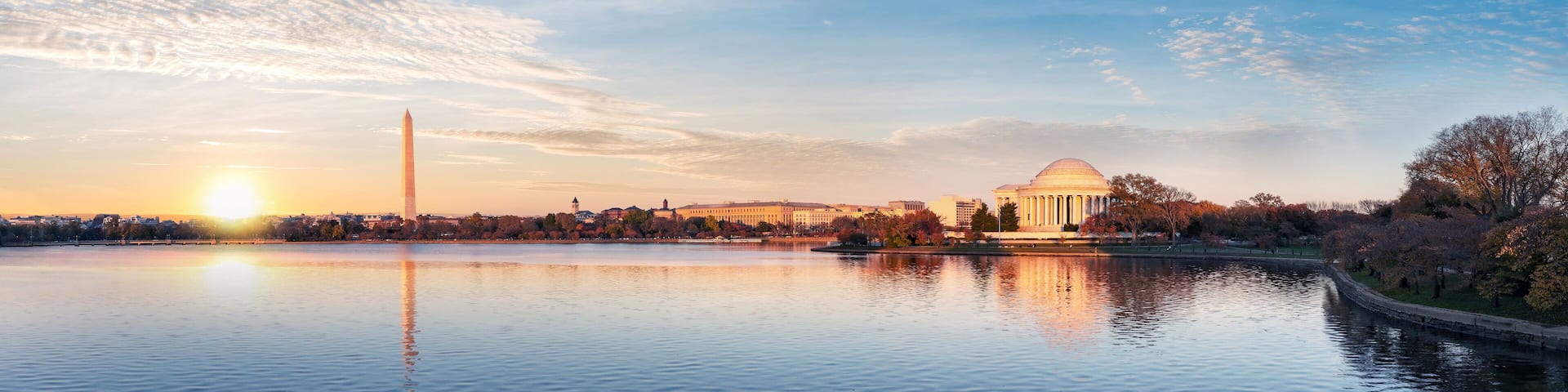 A sunset over a calm lake with a Washington Monument and a domed building reflecting on the water.