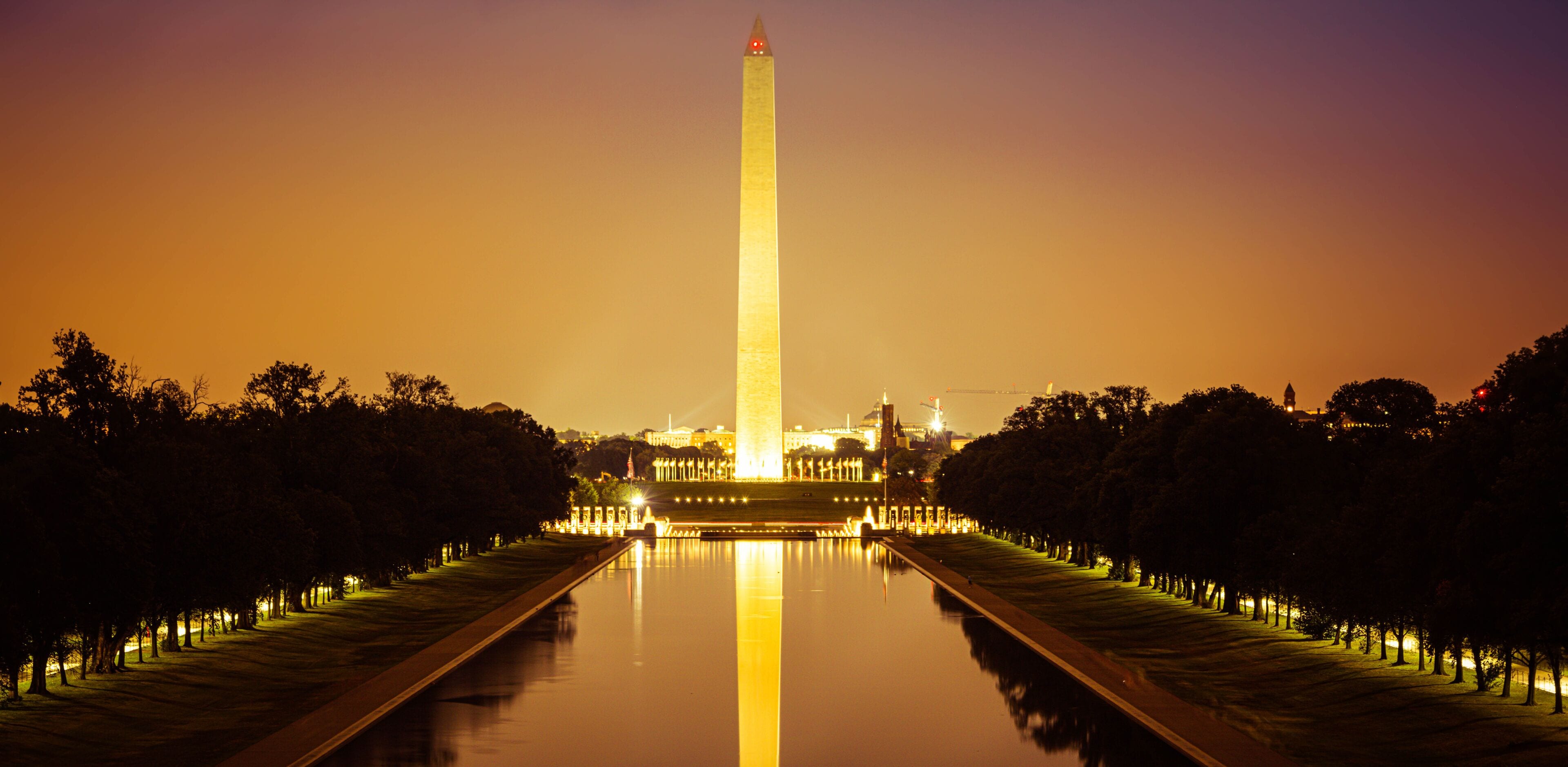 A nighttime view of the Washington Monument with its reflection in the water, surrounded by trees and illuminated pathways.