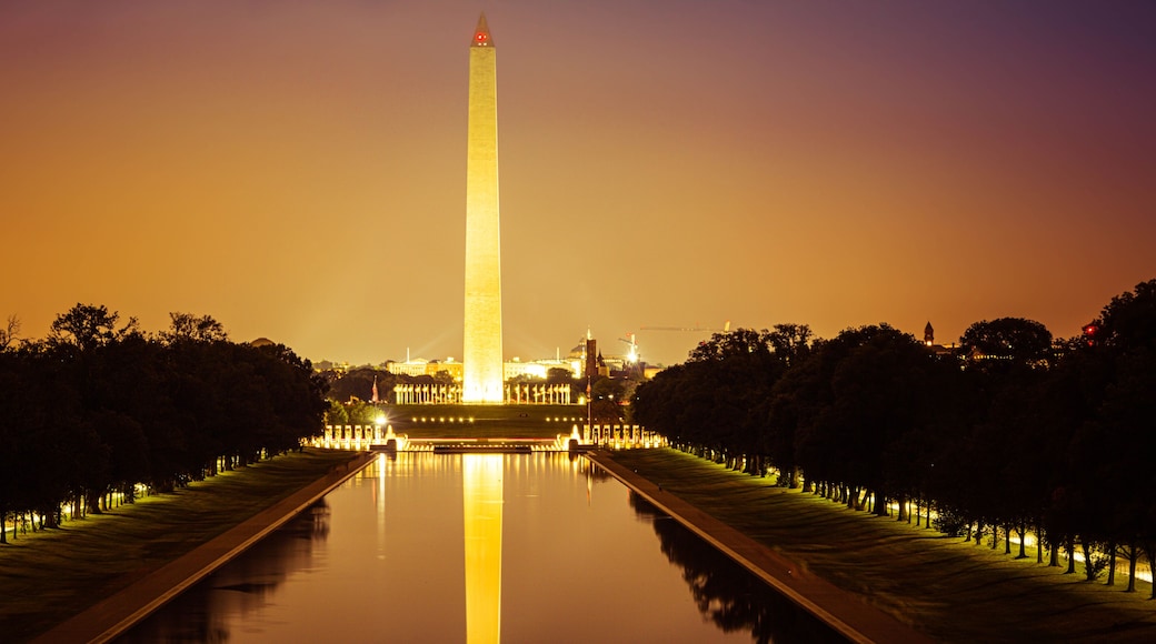 A nighttime view of the Washington Monument with its reflection in the water, surrounded by trees and illuminated pathways.