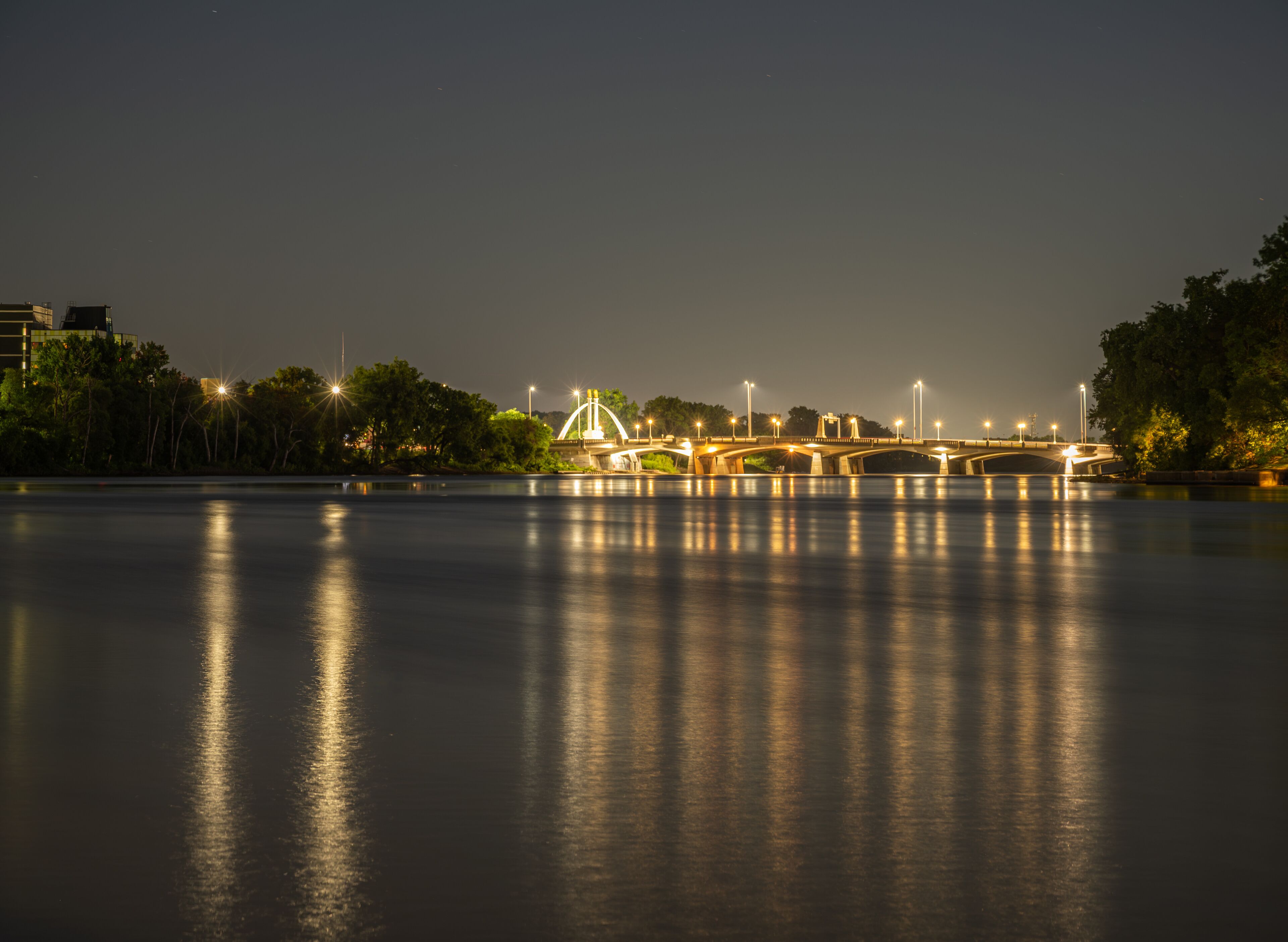 Un río nocturno con un puente iluminado por farolas, reflejado en la superficie del agua.