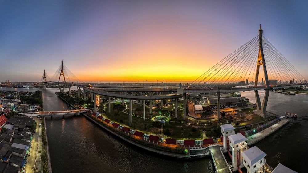 A modern cable-stayed bridge at sunset with a river and traditional buildings nearby.