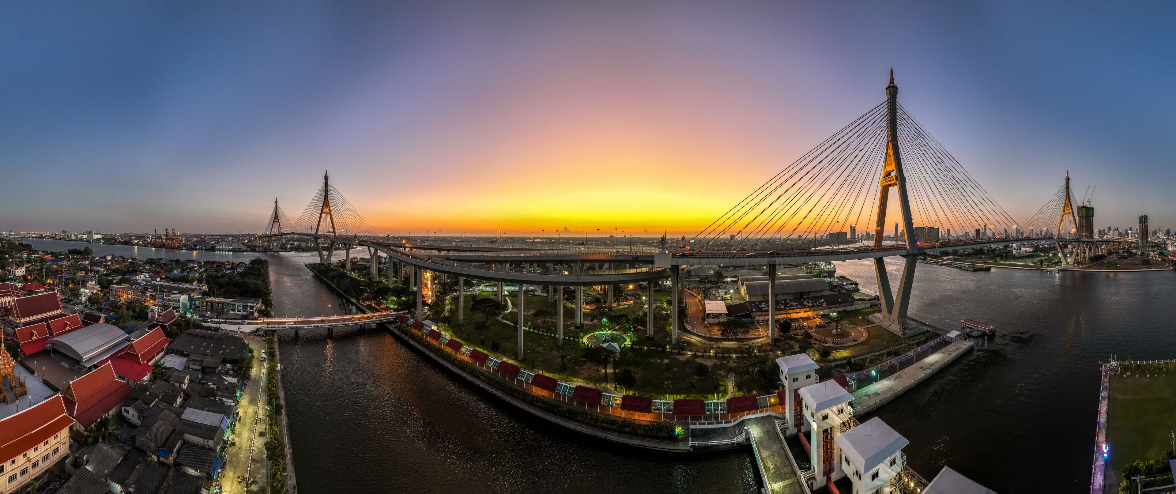 A modern cable-stayed bridge at sunset with a river and traditional buildings nearby.
