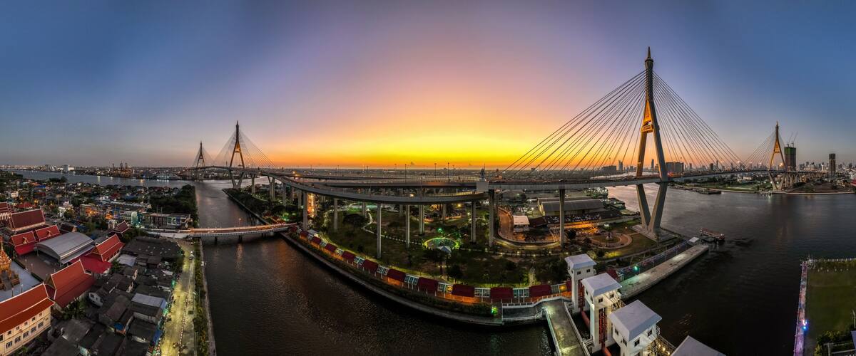 A modern cable-stayed bridge at sunset with a river and traditional buildings nearby.