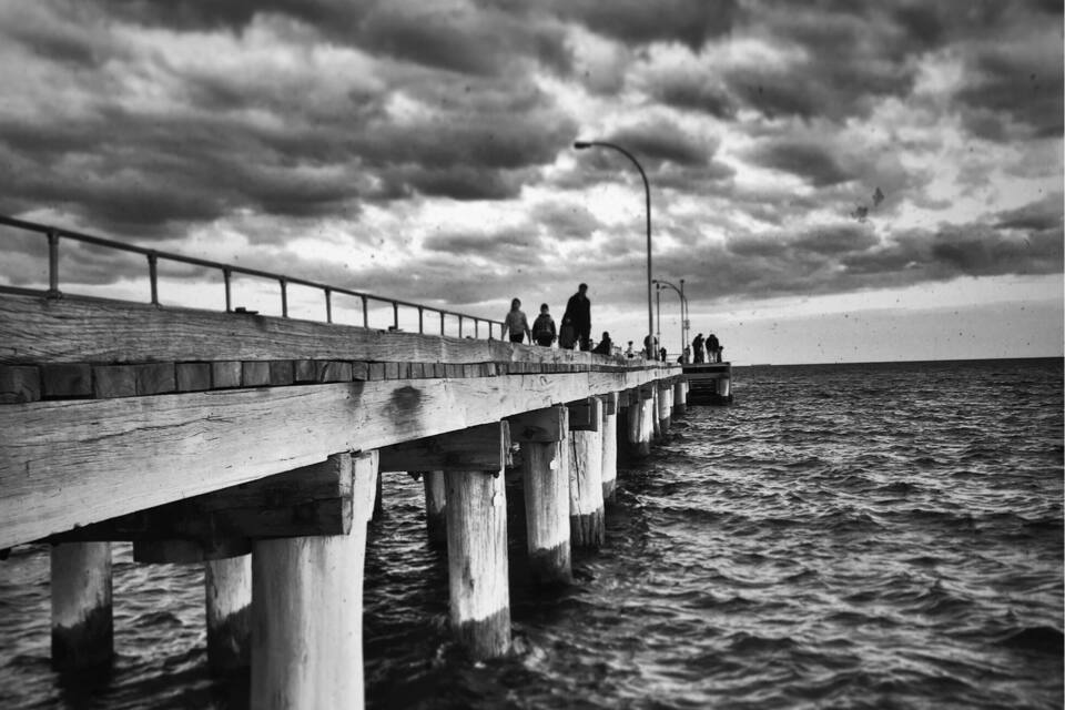 A pier extending into the water with people walking along it under a cloudy sky.