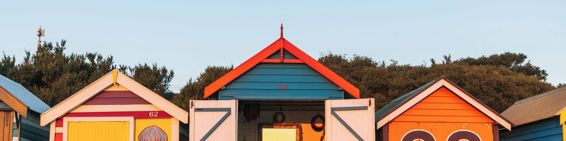 Three colorful beach huts with numbers 62, 63, and 64, and a green kayak in front.
