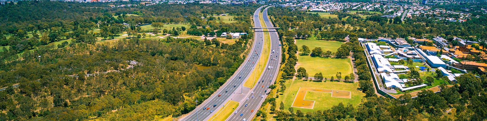 An aerial view of a highway curving through a green landscape with a city skyline in the distance.